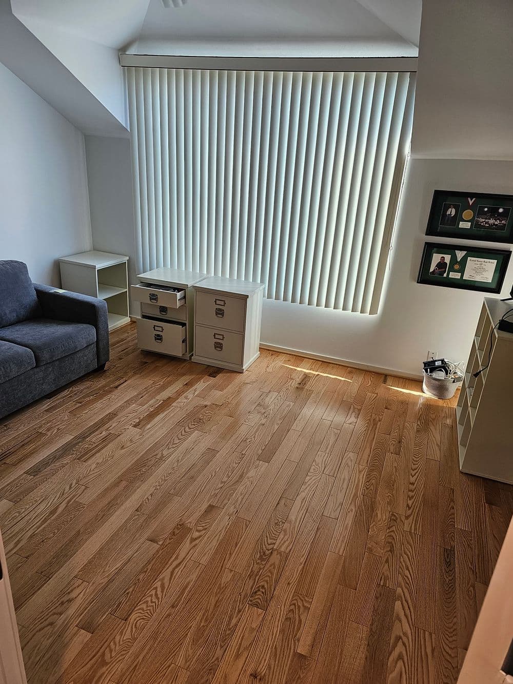 Cozy room with hardwood floor, grey couch, storage cabinets, and large vertical blinds.