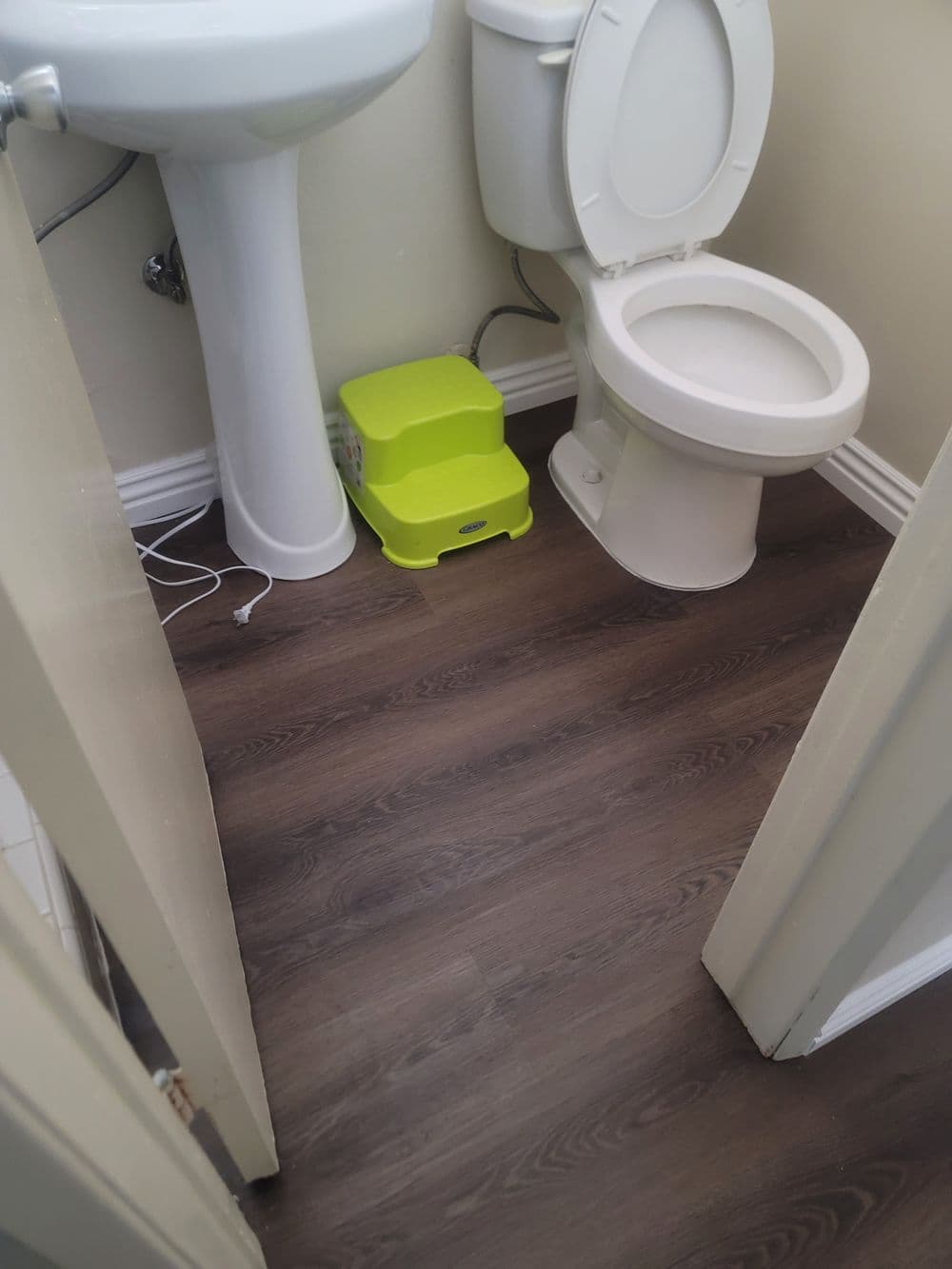 Small bathroom view featuring a toilet, sink, and a green step stool on wood-like flooring.