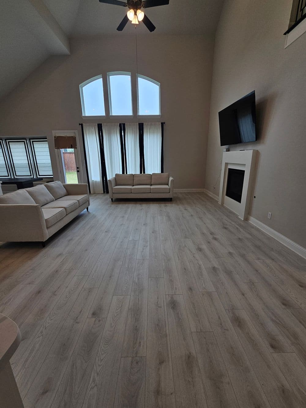 Modern living room with light wood flooring, large windows, beige couch, and wall-mounted TV.