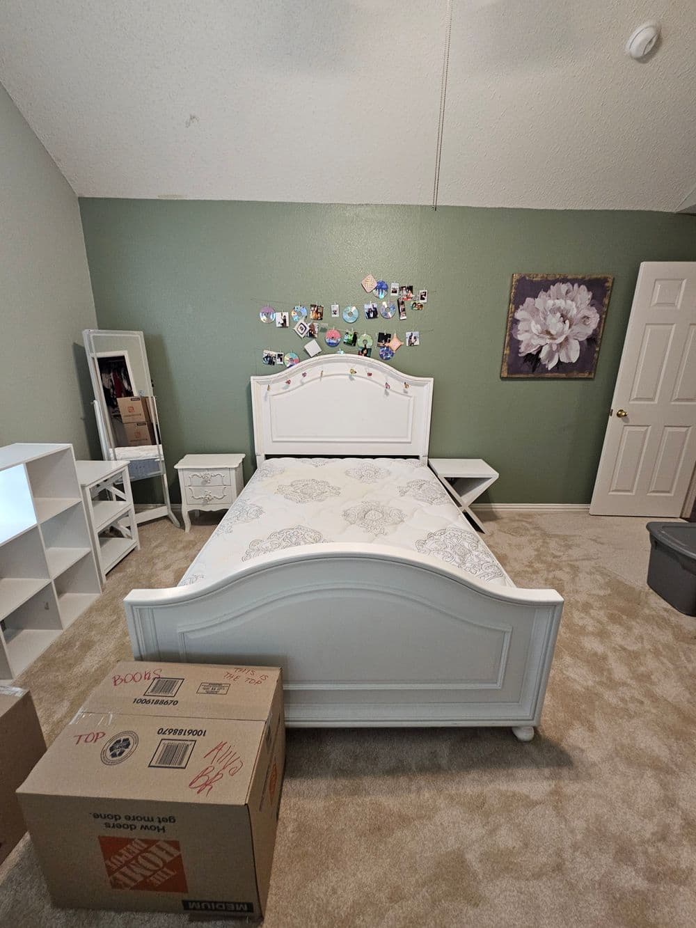 White bed frame with no mattress in green bedroom, featuring a mirror, bookshelf, and moving boxes.