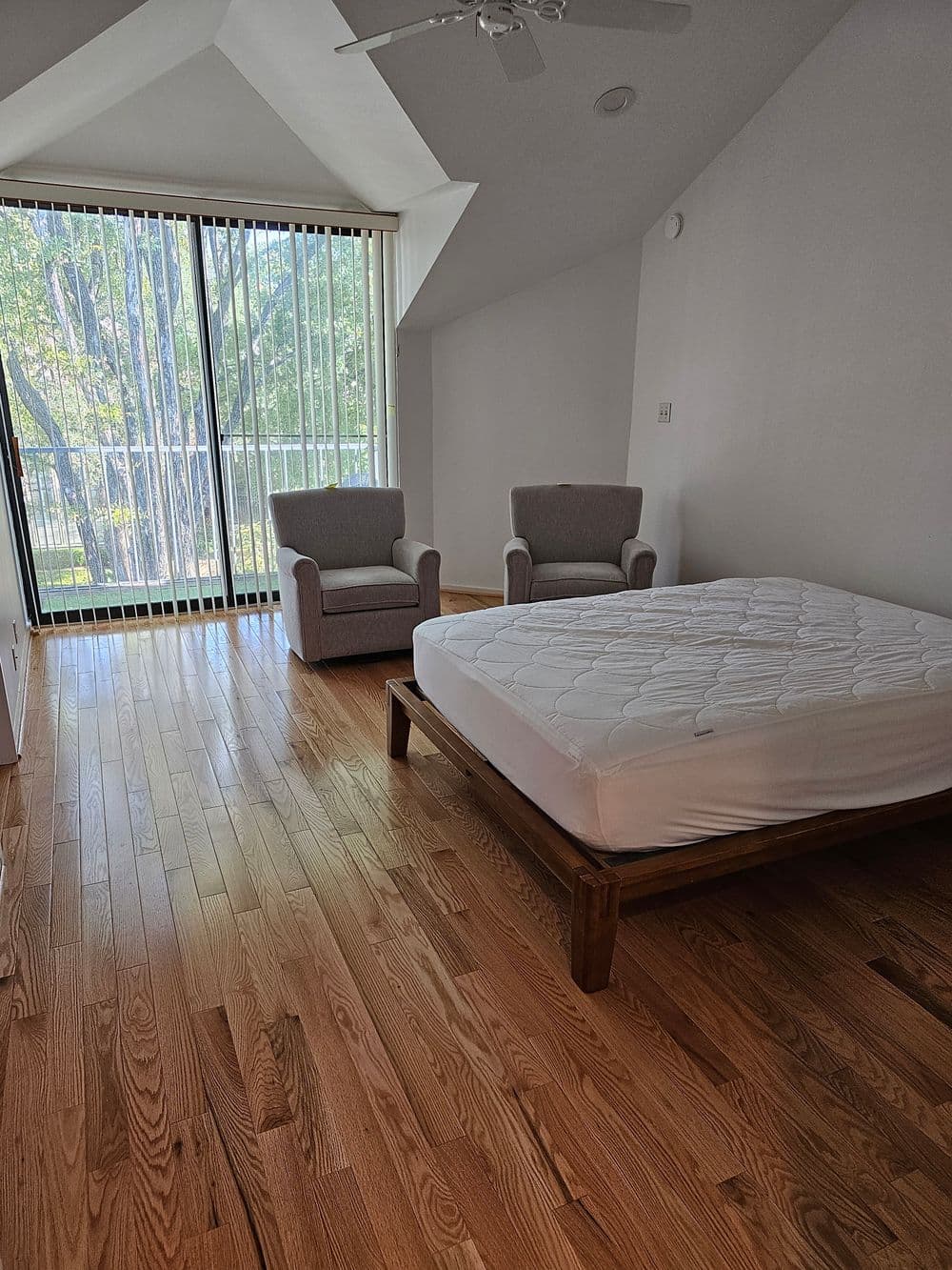 Bright bedroom with hardwood floor, empty mattress on wooden frame, and two chairs by large window.