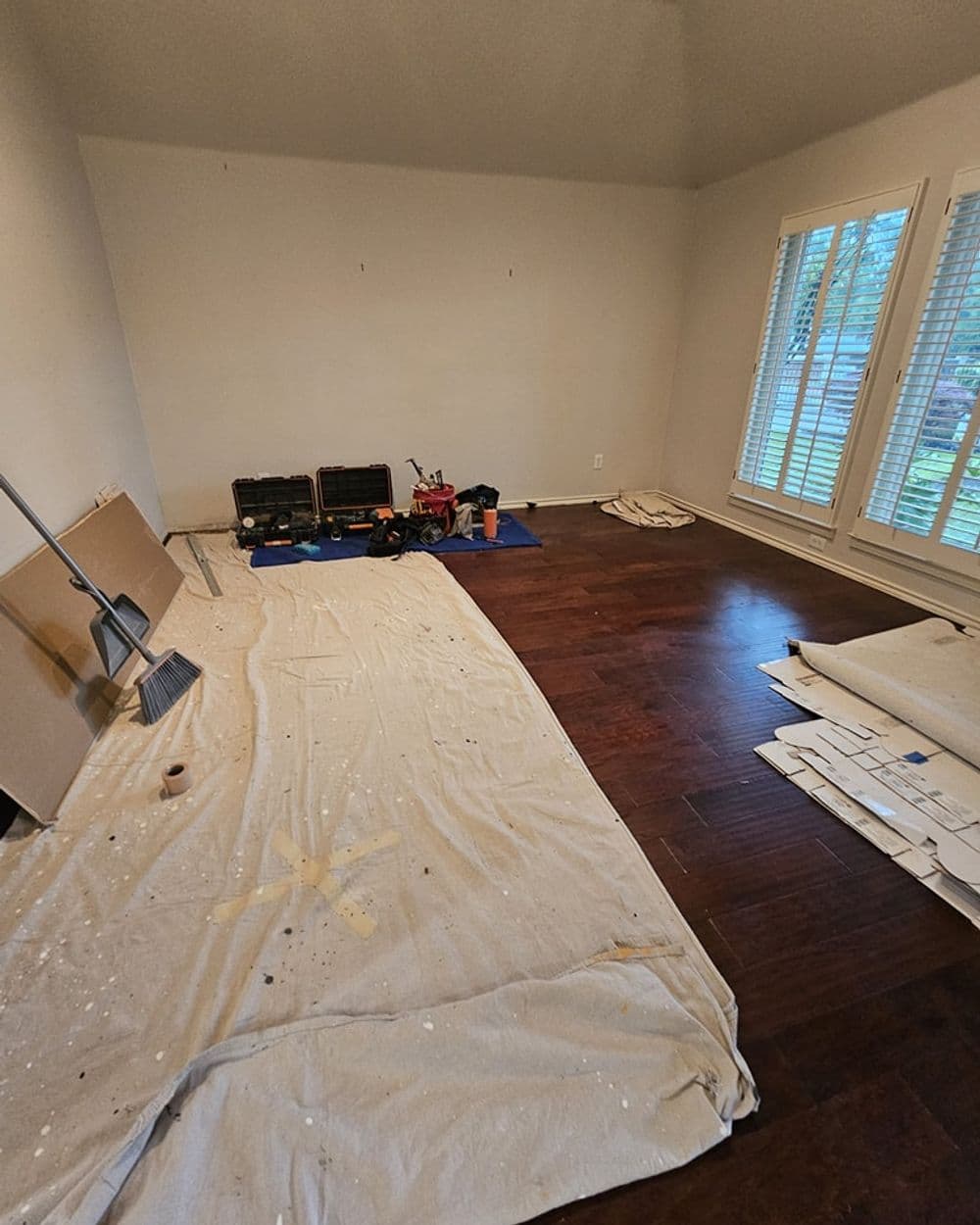 Empty room with hardwood floors covered in painter's tarp and tools for renovation.