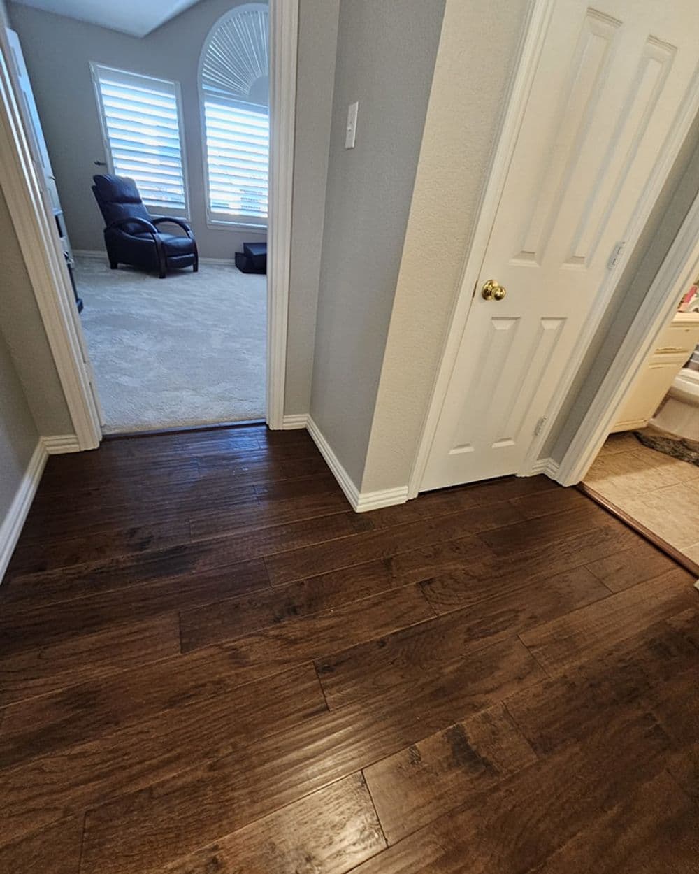Wooden flooring in a hallway leading to a cozy room with a recliner and light-colored carpet.
