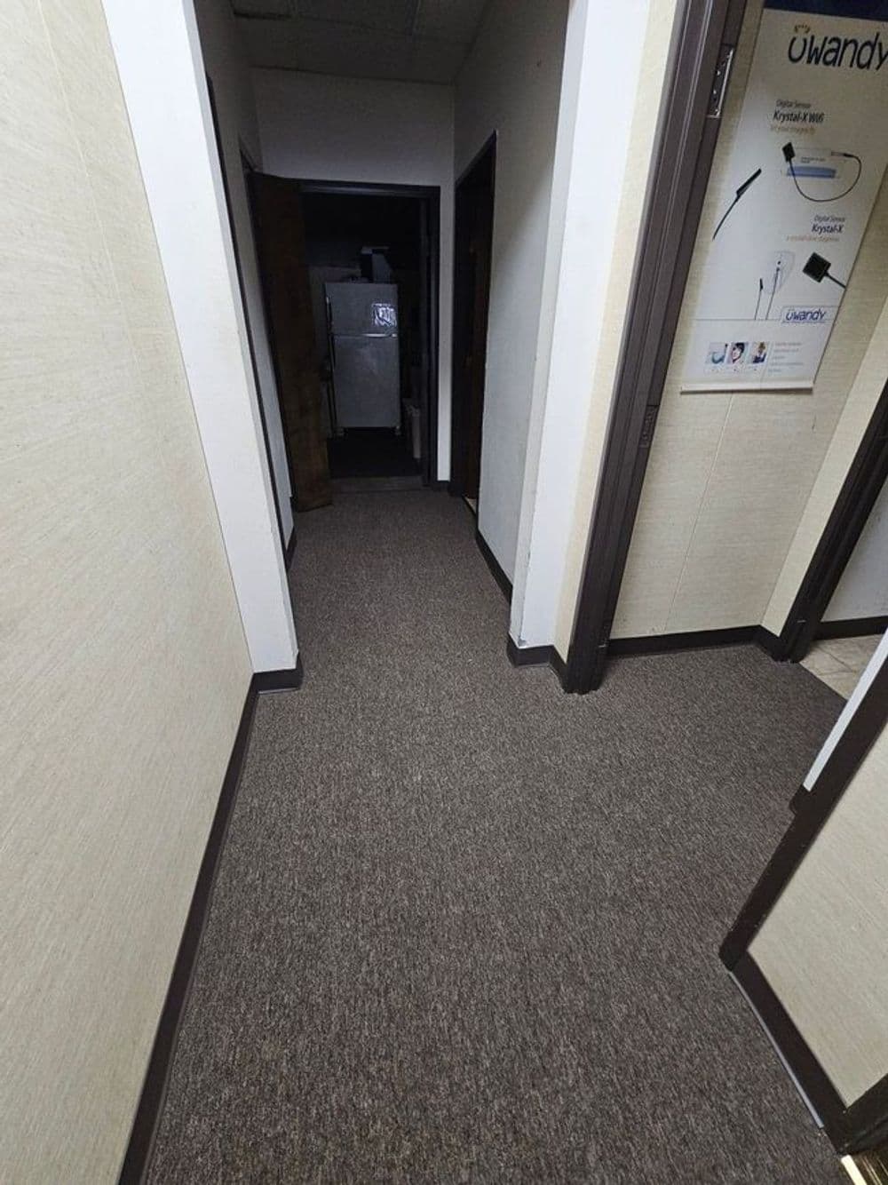 Hallway with beige walls and carpet, featuring open doorways and a dark room at the end.