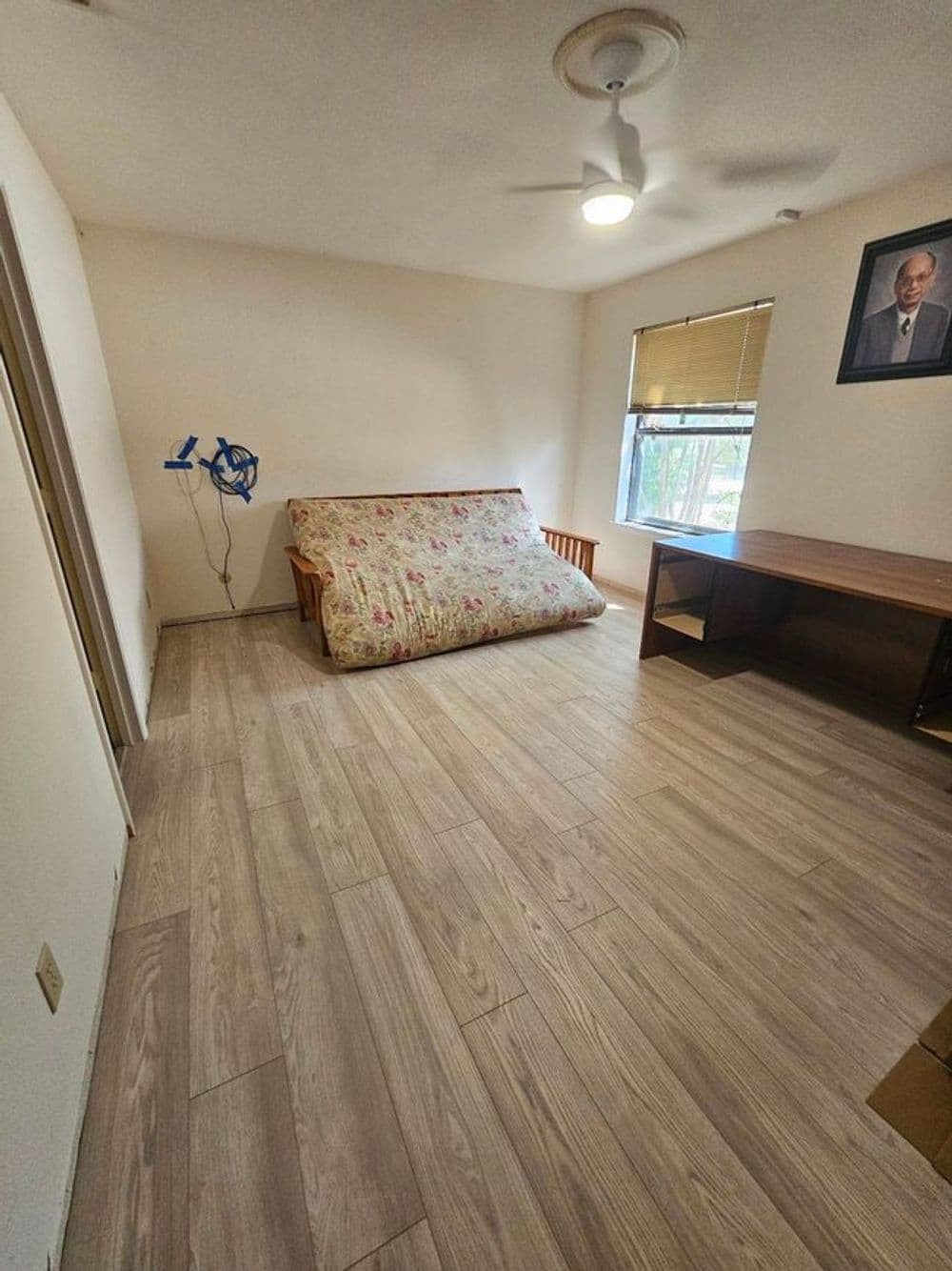 Empty room with wooden flooring, a floral futon, a desk, and a framed portrait on the wall.